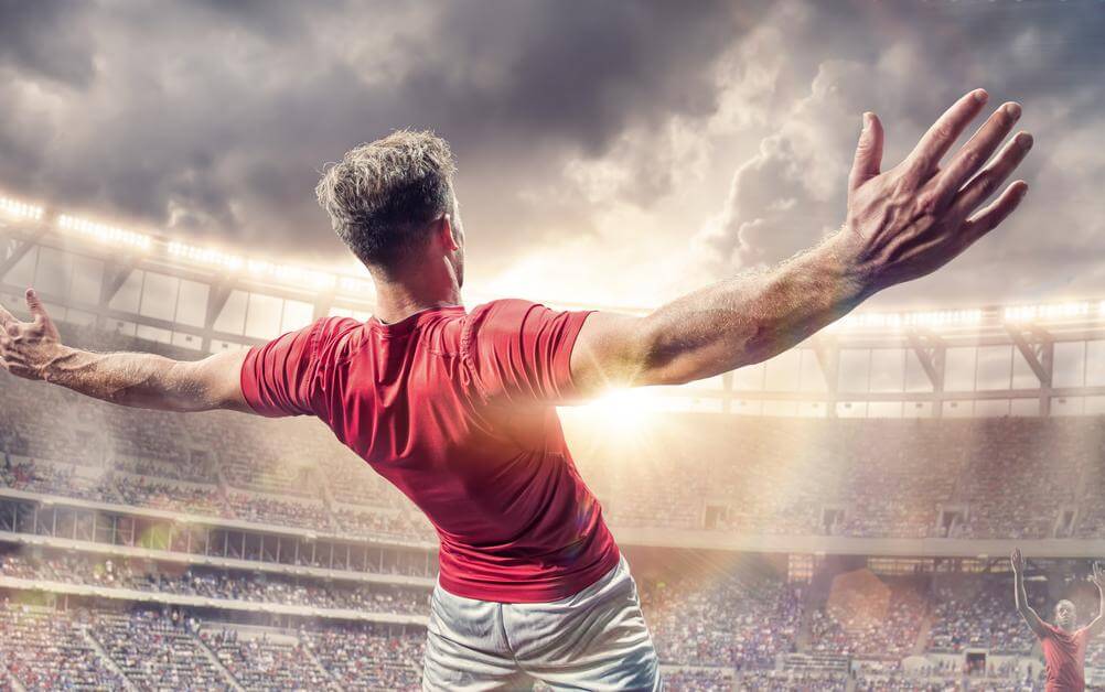 Bild zeigt einen jubelnden Fußballspieler in rotem Trikot mit ausgebreiteten Armen vor einer großen Menschenmenge im Stadion, die sich freuen, dass sie eine "Beide-Teams-Treffen" Wette abgeschlossen haben.